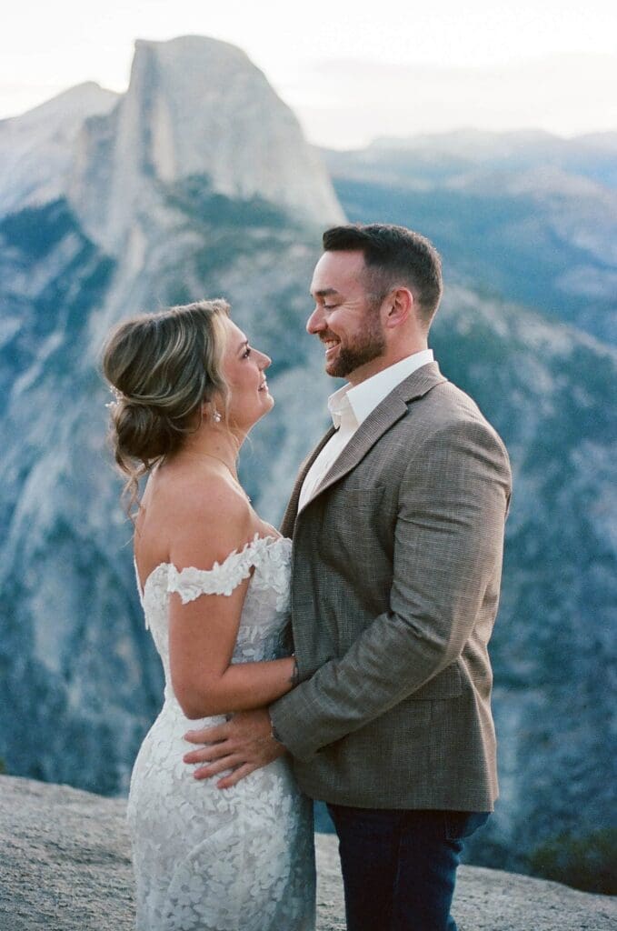 Captured on 35mm film, a bride and groom stand in a face to face embrace in the shadow of the enormous Half Dome granite monolith at Glacier Point in Yosemite National Park, California.