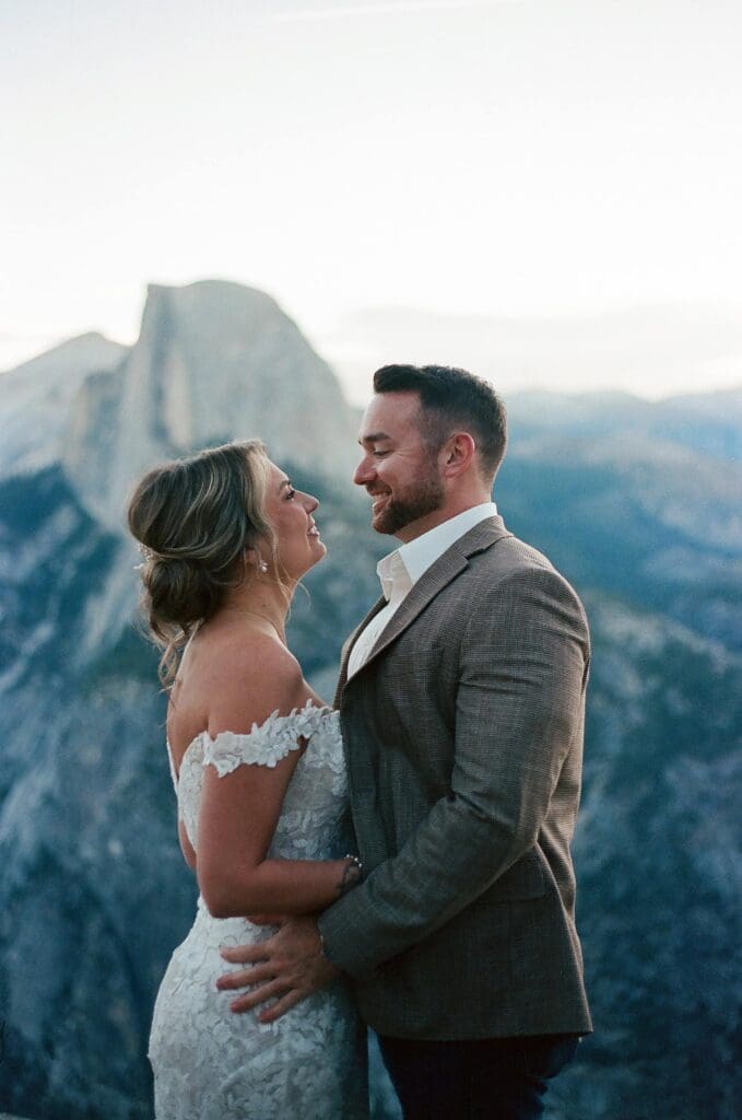 Captured on 35mm film, a bride and groom stand in a face to face embrace in the shadow of the enormous Half Dome granite monolith at Glacier Point in Yosemite National Park, California. 