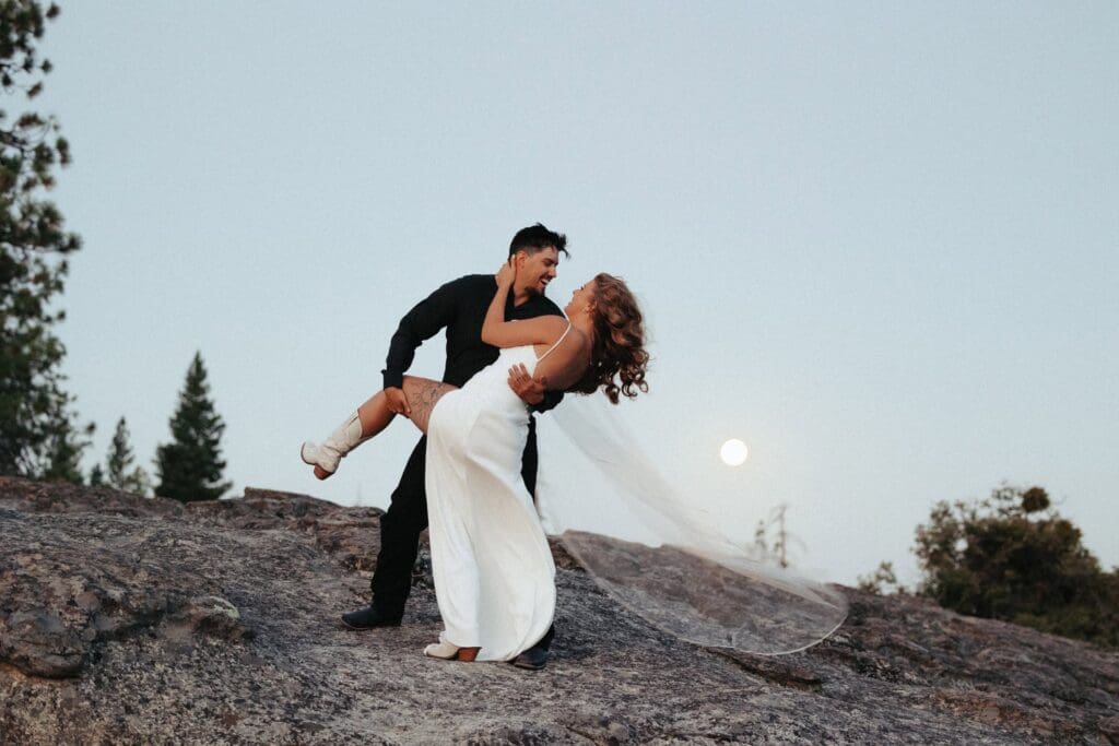 A groom dips his bride atop a swath of exposed granite bathed in moonlight of the setting full moon shining through the bride's veil. 