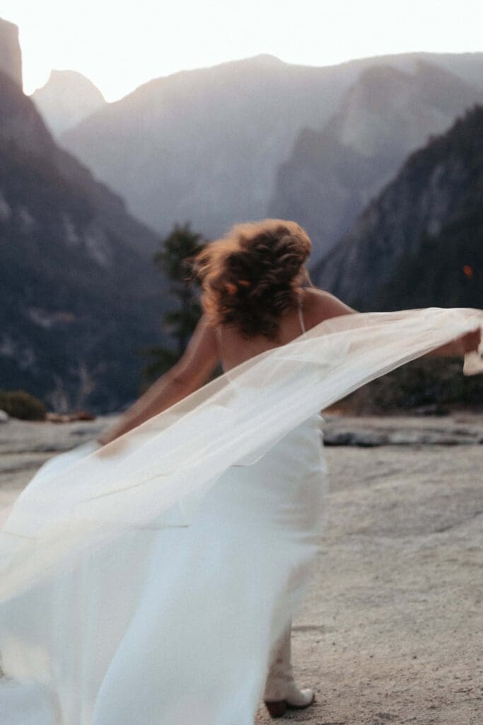 A bride runs, hair flowing and veil trailing behind her, blurred by motion and morning light, across a stretch of exposed granite at Yosemite National Park's Tunnel View. Half Dome and El Capitan loom on the horizon, lit by dawn's first light. 