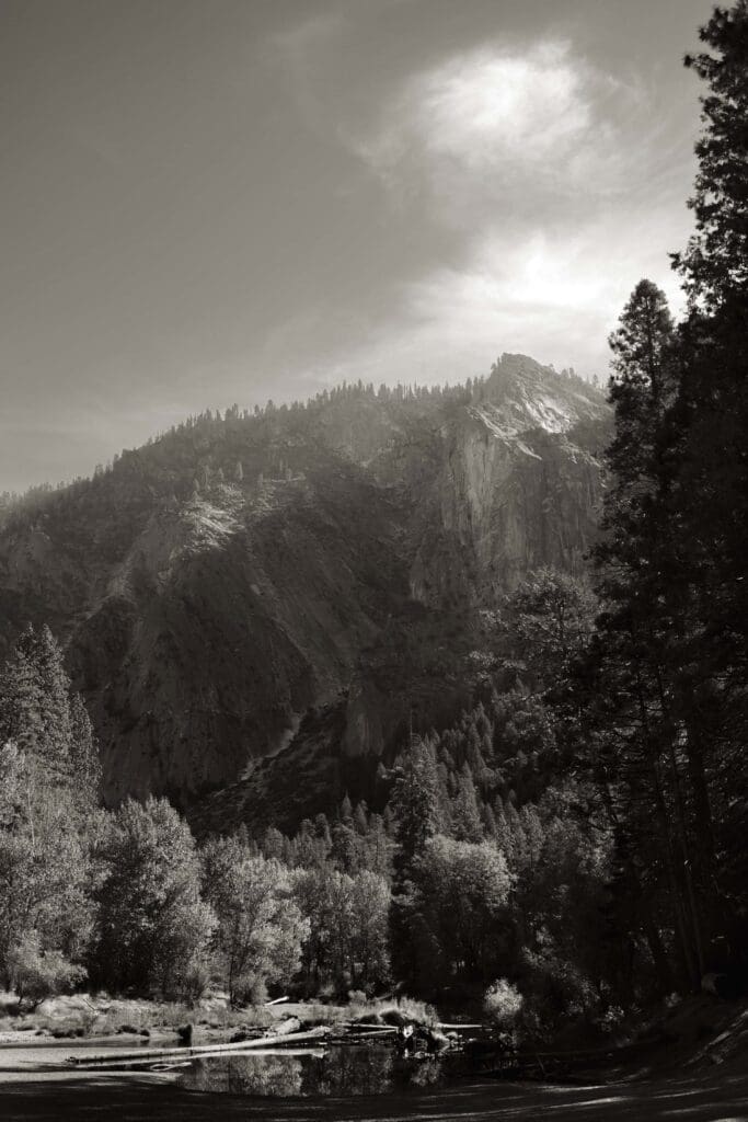 A black and white image of an alpine beach with mountains and pines and interesting light pouring out of a partly cloudy sky.