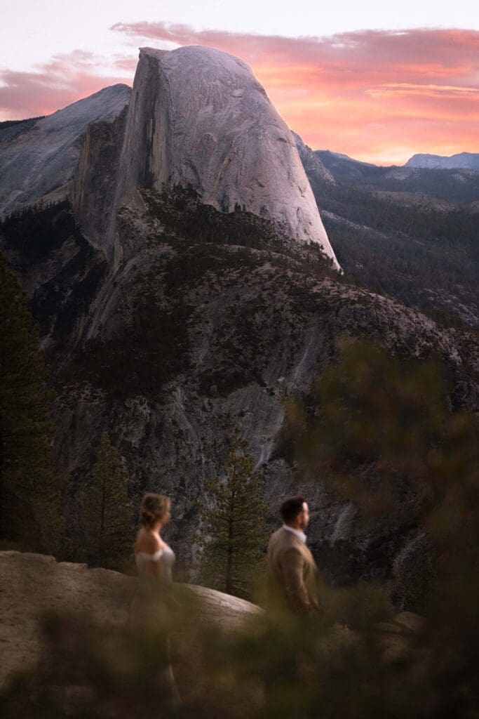 A bride faces her groom as his back is turned in the moments before their first look. Partially obscured by flora in the foreground, Half Dome looms behind in the dawn light.