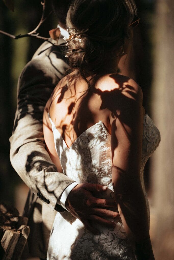 A bride and groom, pictured up close and at an angle from the back, looking into a forest of redwoods at the Mariposa Grove as the shadows of leaves dance across the bride's back above the plunging backline of her gown. 