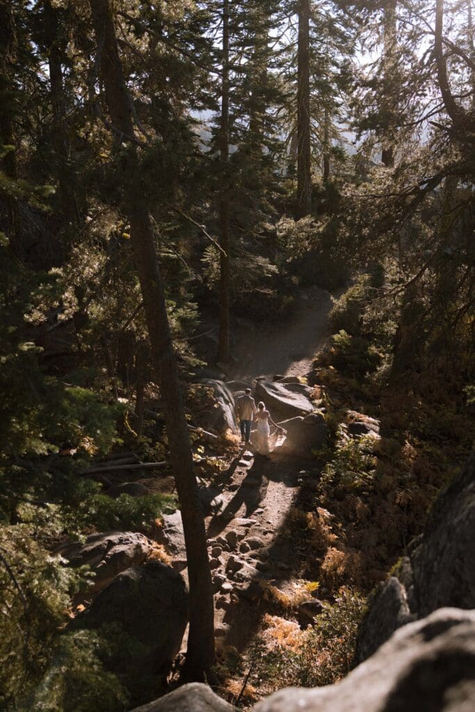 A bride and groom walk hand in hand over a rocky trail through a glowing sunlit forest of pine trees and granite boulders approaching Yosemite National Park's iconic Taft Point view.