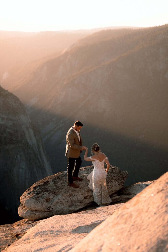 A groom extends a hand to his bride as she steps onto a sparkling granite boulder overlooking sweeping views of a golden-hour Yosemite Valley at Taft Point.
