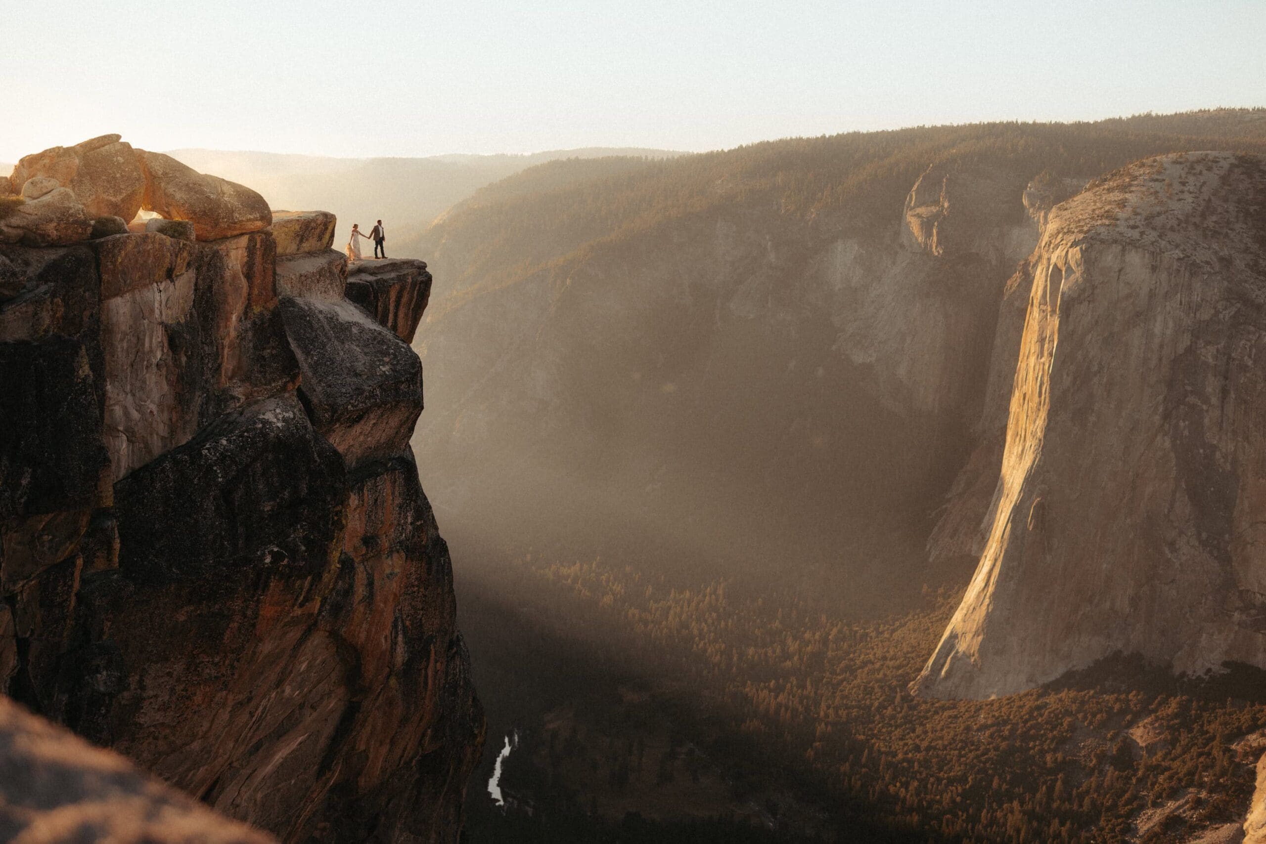 A bride and groom stand atop Taft Point at a dizzying height, overlooking Yosemite Valley and El Capitan drenched in golden-hour light in Yosemite National Park, California.