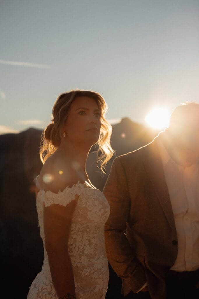 A bride and groom stand before a Yosemite sunrise, sun flare grazing the groom's shoulder as he looks down and the bride's neckline as she looks off into the distance atop Glacier Point.