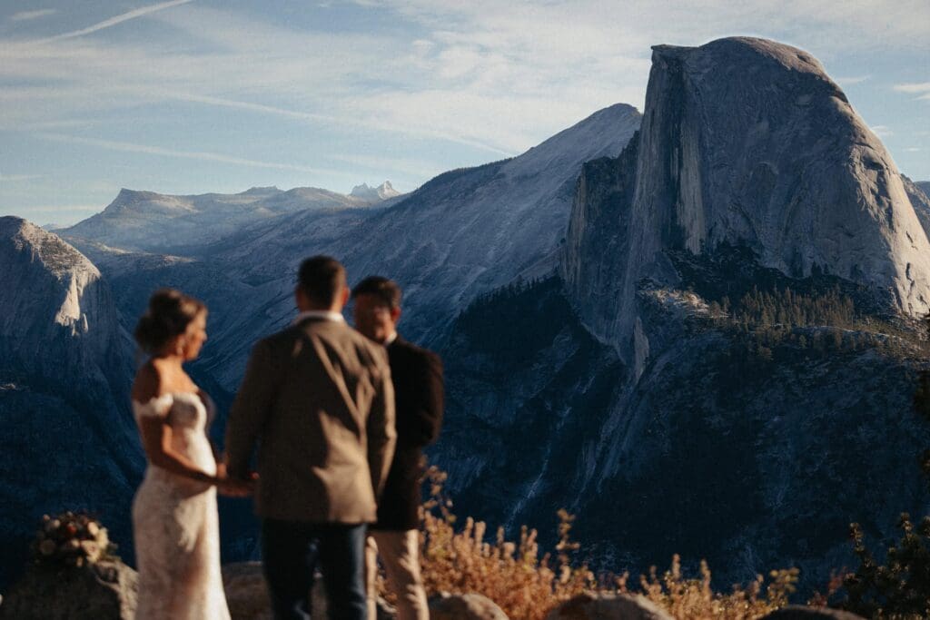 A bride and groom stand before a minister, out of focus, while Half Dome looms gracefully above them, in perfect focus and vivid alpine blue color in the morning light.