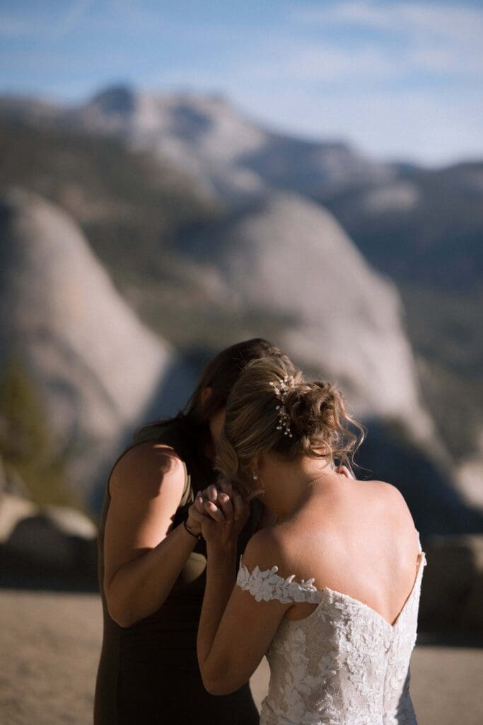 A bride leans forehead to forehead with her sister, while clasping hands and poised with excitement and emotion atop Glacier Point at Yosemite National Park, California. 